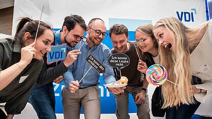 Sechs Young Engineers stehen um einen Teller mit Gebäck und halten humorvolle Schilder sowie Requisiten wie ein buntes Lollipop.