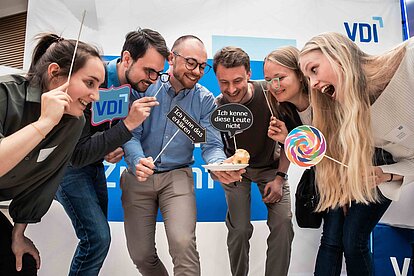 Sechs Young Engineers stehen um einen Teller mit Gebäck und halten humorvolle Schilder sowie Requisiten wie ein buntes Lollipop.