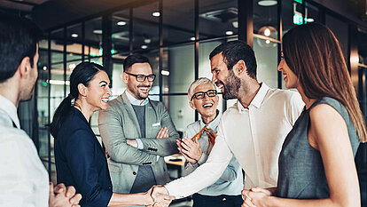 Sechs Personen stehen in einem modernen Büro. Zwei in der Mitte geben sich die Hand, die anderen schauen lächelnd zu.