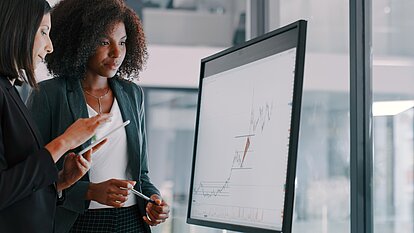 Zwei Personen stehen vor einem großen Monitor. Auf dem Monitor ist ein Diagramm mit einer ansteigenden Kurve und mehreren horizontalen Linien zu sehen.