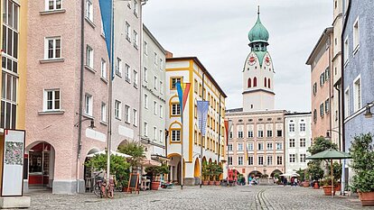 Belebter Platz mit Kopfsteinpflaster in Rosenheim. Umgeben von pastellfarbenen Gebäuden mit Balkonen und Cafés. In der Mitte steht ein Turm mit grüner Kuppel und Uhr.