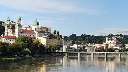 Panorama von Passau mit der Donau im Vordergrund, historischen Gebäuden und dem Dom St. Stephan. Eine Brücke verbindet die Ufer, im Hintergrund bewaldete Hügel.